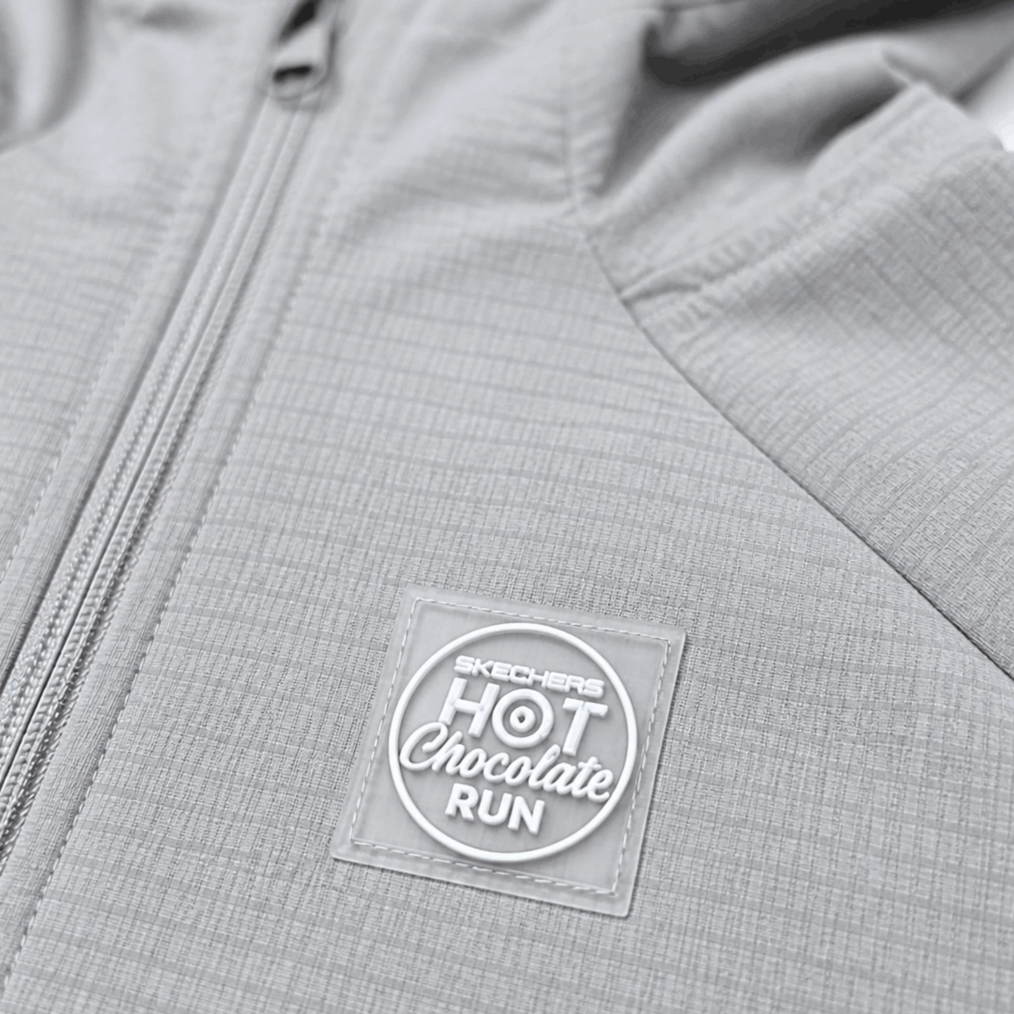 Close-up of a light gray zippered jacket with a circular Skechers Hot Chocolate Run rubber stamp