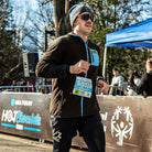 A man running wearing a brown Hot Chocolate Run fleece zip-up hoodie with bright blue accents. He has a race bib and standing in front of a race barrier with 'Hot Chocolate Run' branding