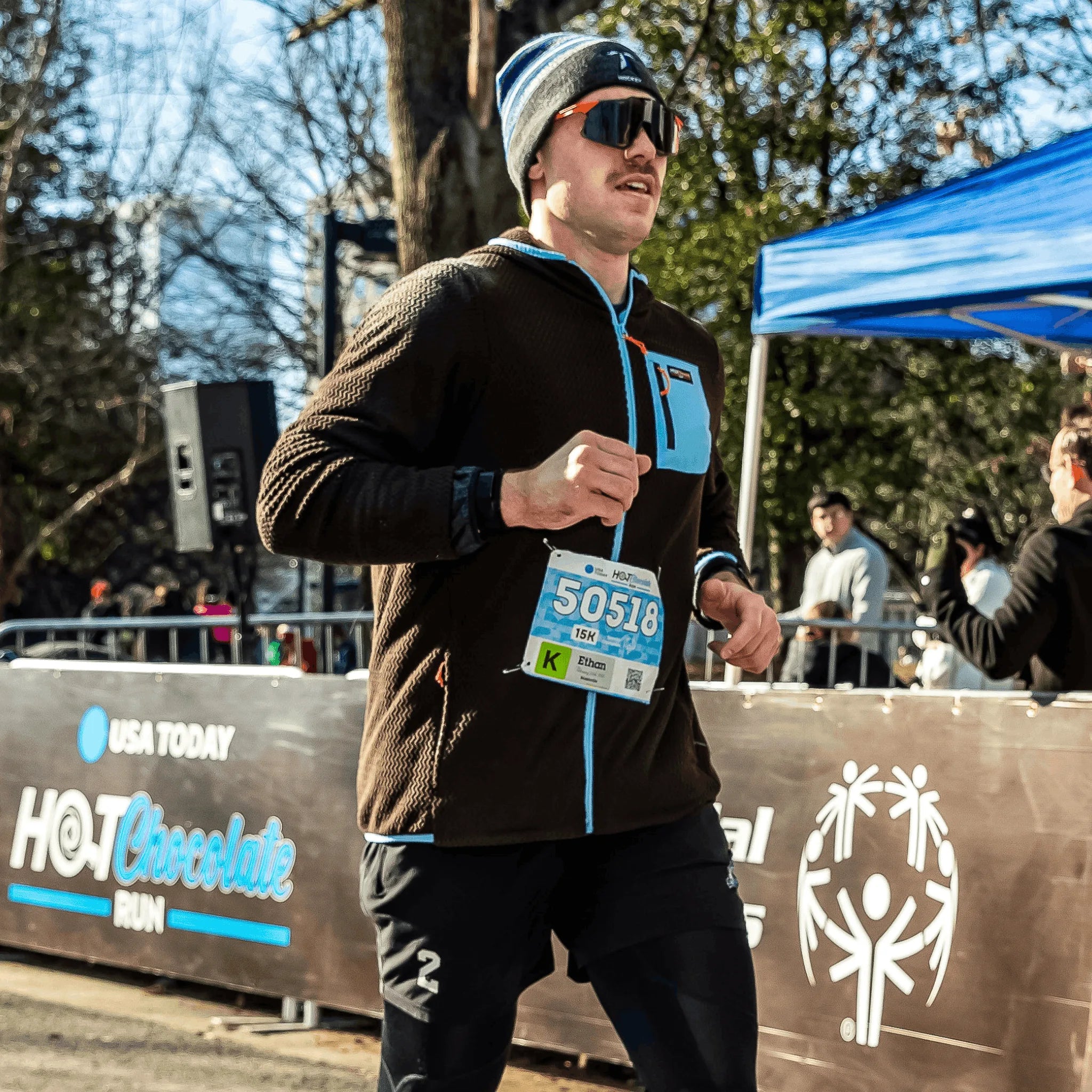 A man running wearing a brown Hot Chocolate Run fleece zip-up hoodie with bright blue accents. He has a race bib and standing in front of a race barrier with 'Hot Chocolate Run' branding