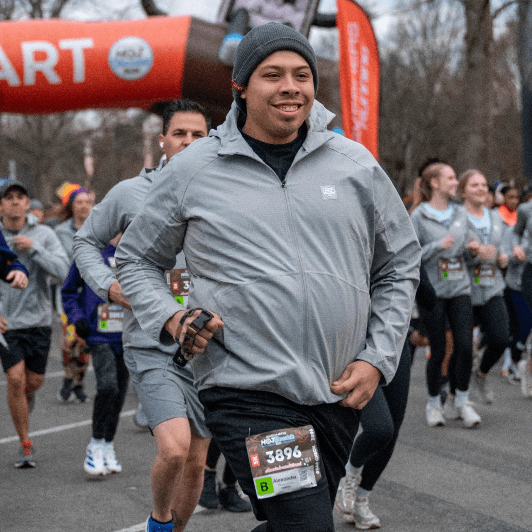 A man running wearing a light gray Hot Chocolate Run zip-up jacket with a race bib pinned to his shorts.