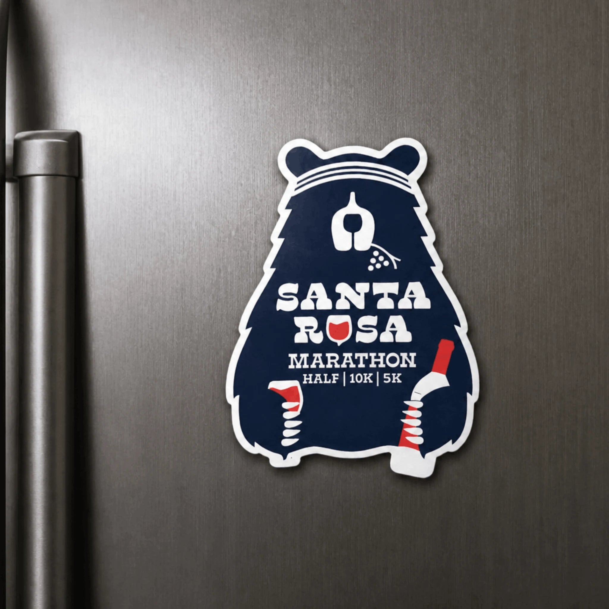 A Santa Rosa Marathon magnet shaped like a bear, featuring a dark navy blue background with white and red text and graphics, shown applied to a dark metallic refrigerator door