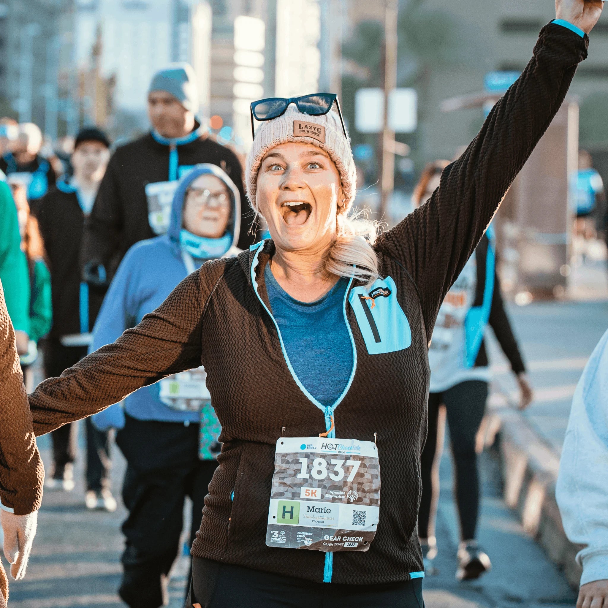 A woman running with her arms raised in celebration, wearing a brown Hot chocolate fleece zip-up hoodie over a blue shirt and a race bib.