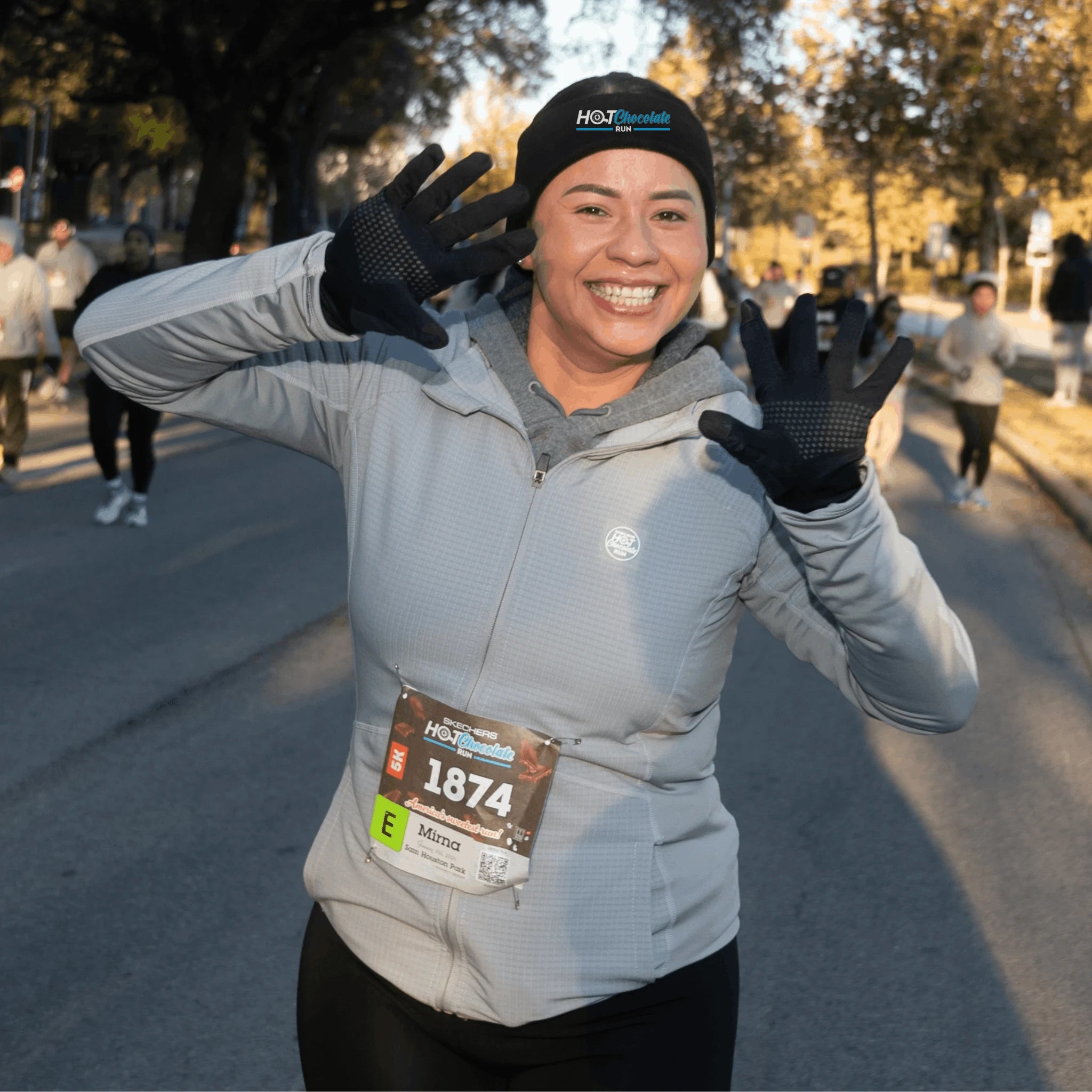 A smiling woman wearing a light gray Hot Chocolate Run zip-up jacket, black headband and gloves poses with her hands raised in front of her face 